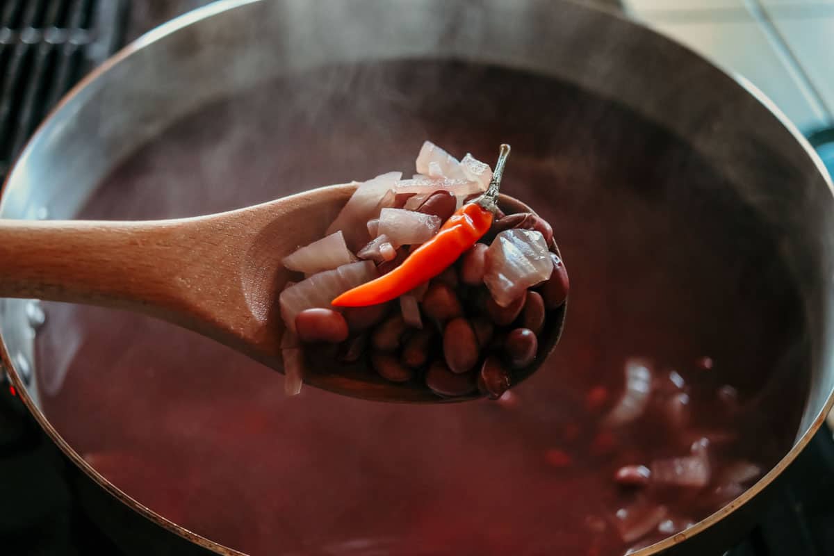 closeup of a bird's eye chile, domingo rojo beans, onions, and other ingredients on a wooden spoon with a red bean broth and more ingredients in a saucier in the background