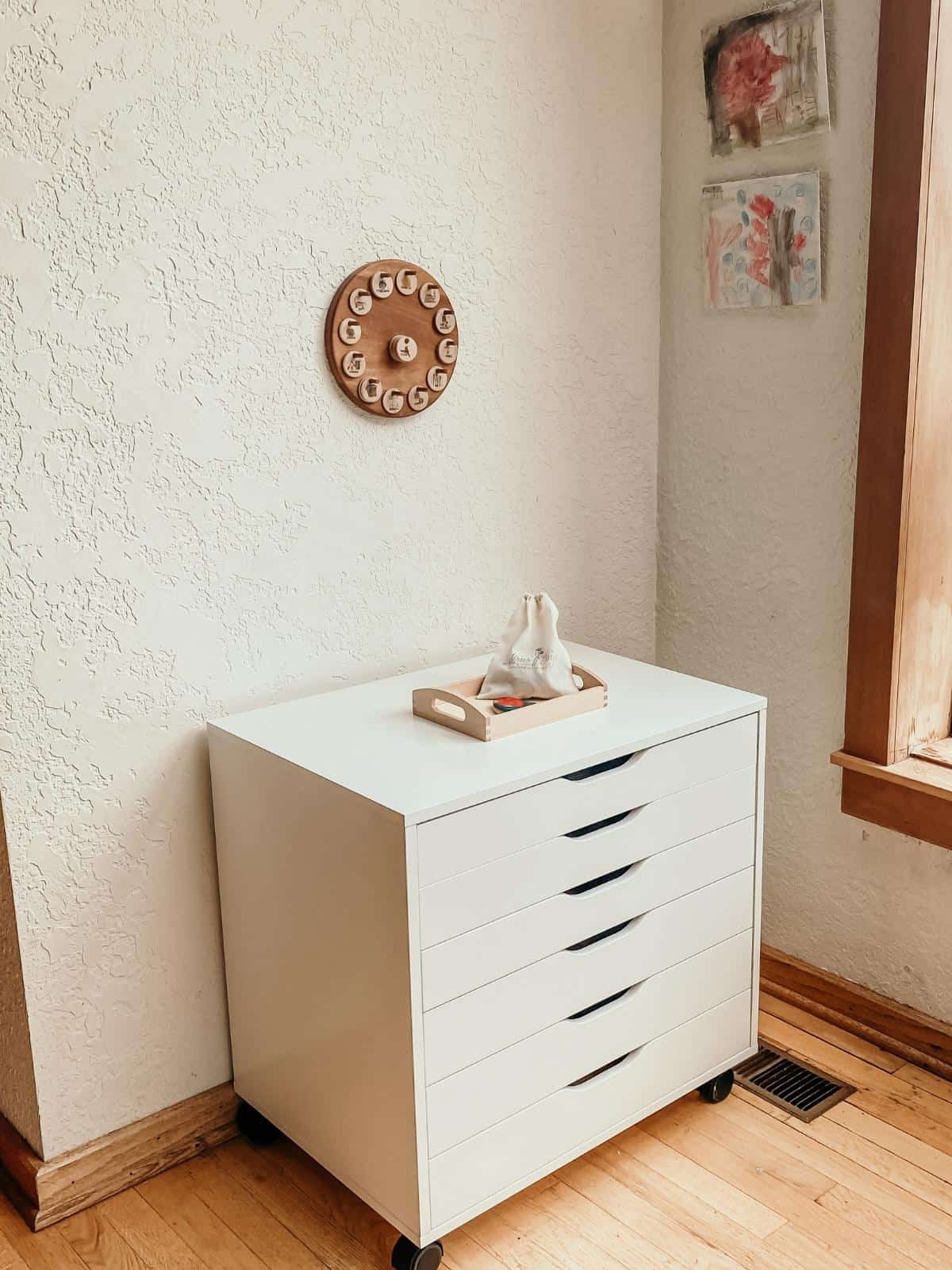 an art cabinet with art activity tray on top in a corner of a home.  There is a wooden daily schedule and watercolor paintings on canvas hanging on the wall.