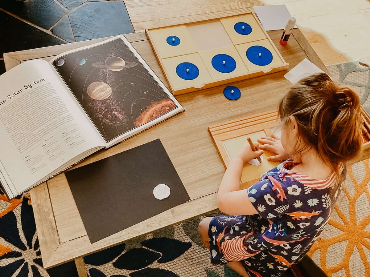 Montessori First Great Lesson Follow-up work:  child tracing Geometry Cabinet Circles Drawer and Planetarium book open to The Solar System