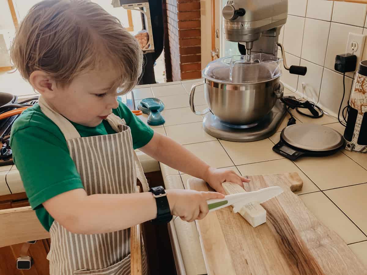 child standing in a learning tower in the kitchen and cutting butter with a child-friendly knife
