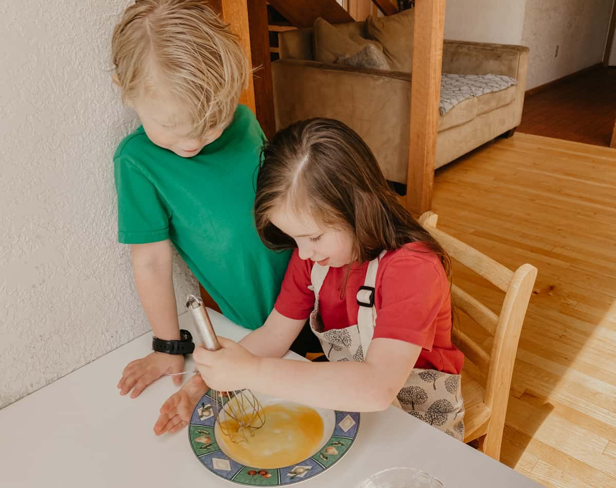 two kids at a table.  one child is whisking eggs in a bowl with a whisk
