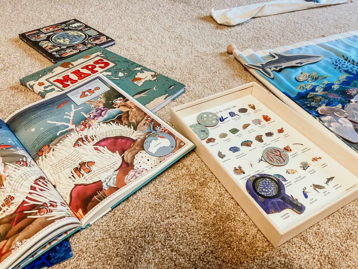 Atlas of Ocean Adventures open to a section about The Great Barrier Reef, Maps book, and The Wondrous Workings of Planet Earth on a rug next to the Waseca Biomes Coral Reef Habitat Mat materials.
