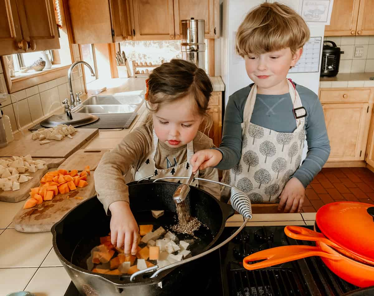 child placing cubes of butternut squash and sweet potato to a dutch oven.  Another child is adding anise seeds to the dutch oven.