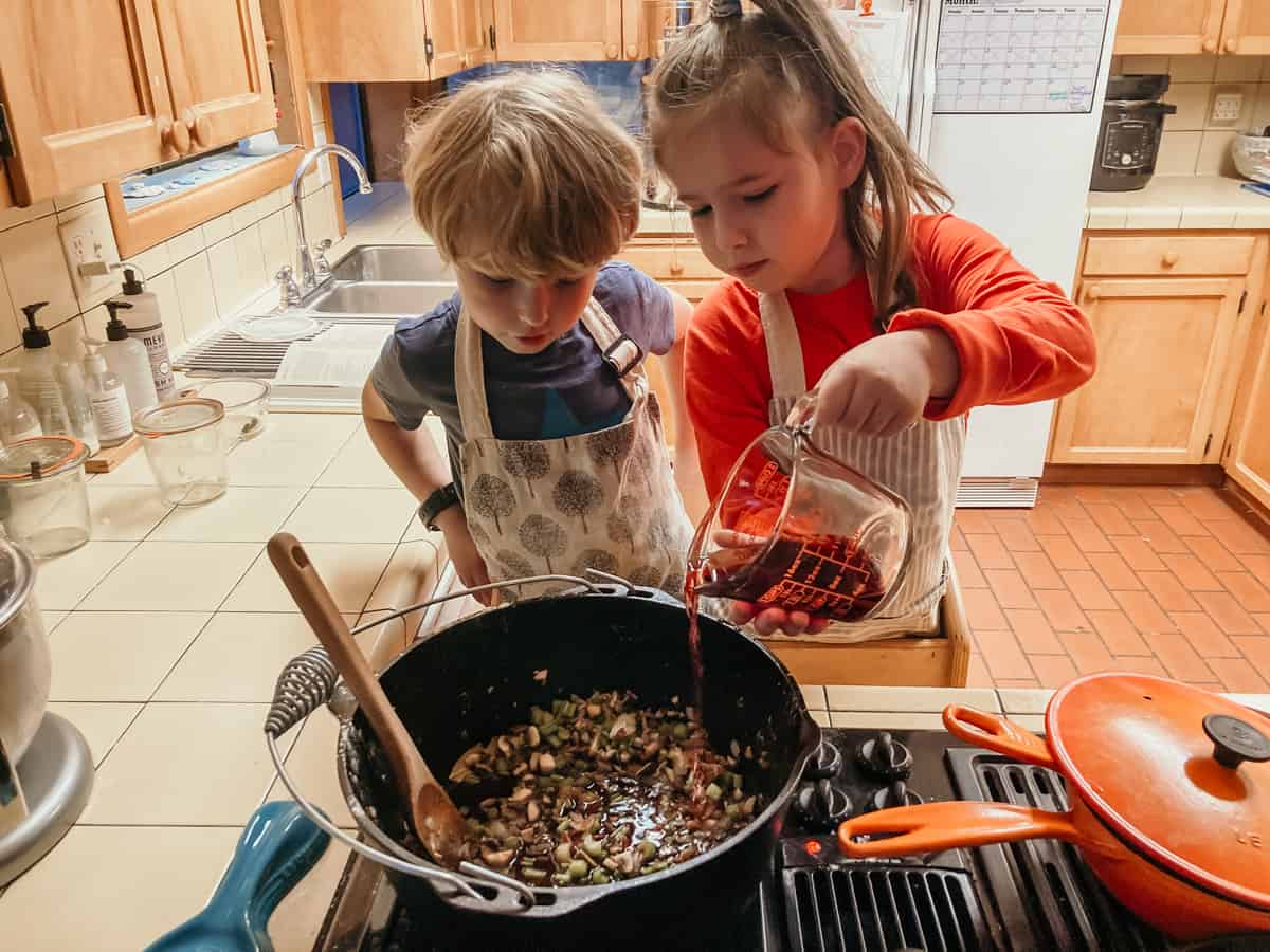 two kids at a stove in a learning tower.  One child is pouring red wine into a dutch oven filled with vegetables.  The other child is watching.