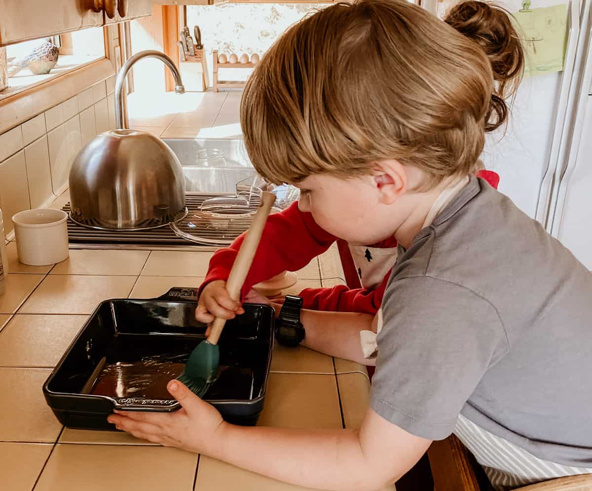one child is holding a baking dish and another child is using a basting brush to apply butter to the dish