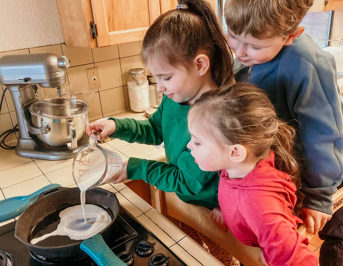 3 kids in a learning tower. one child is pouring milk into a cast iron skillet using a measuring cup.