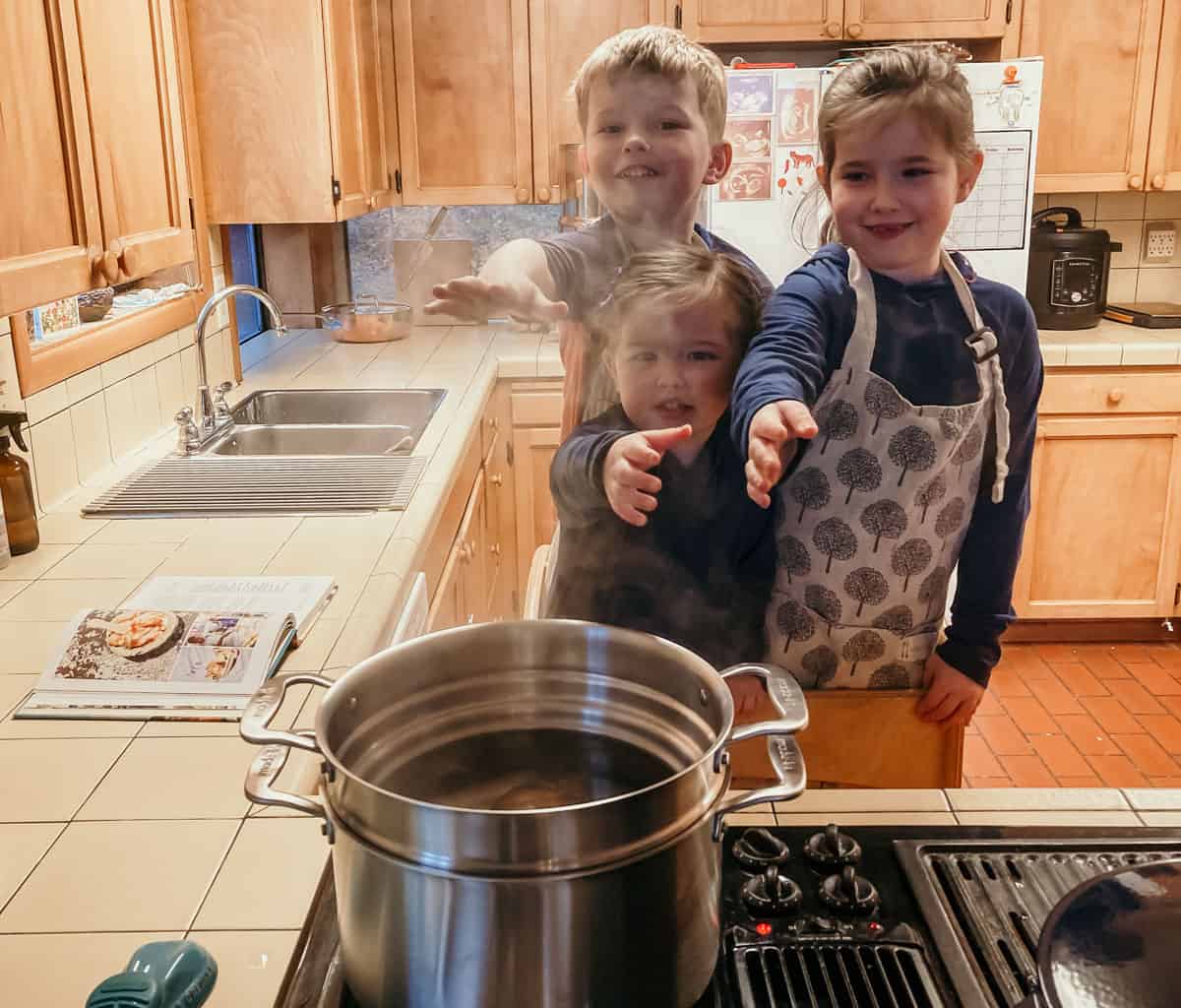 kids standing at the stove in a learning tower with their hands extended over a stock pot