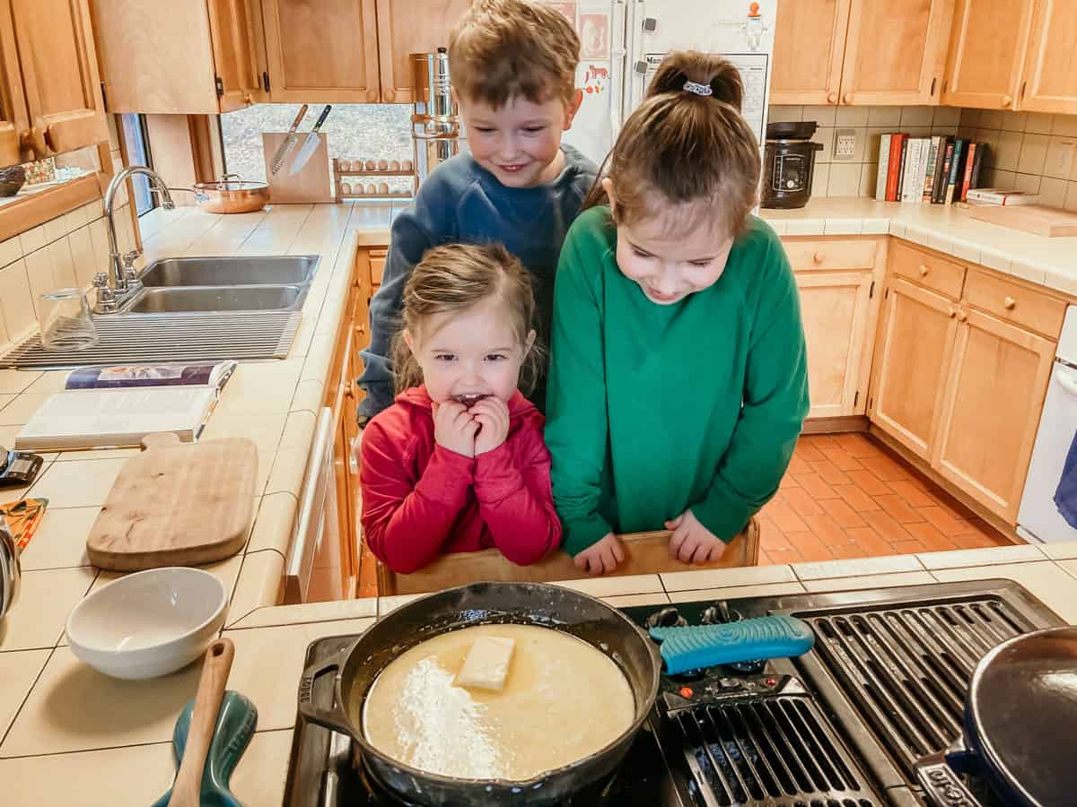 3 kids in a learning tower at the stove. there's a pan on the stove and the kids are waiting for the butter inside to melt