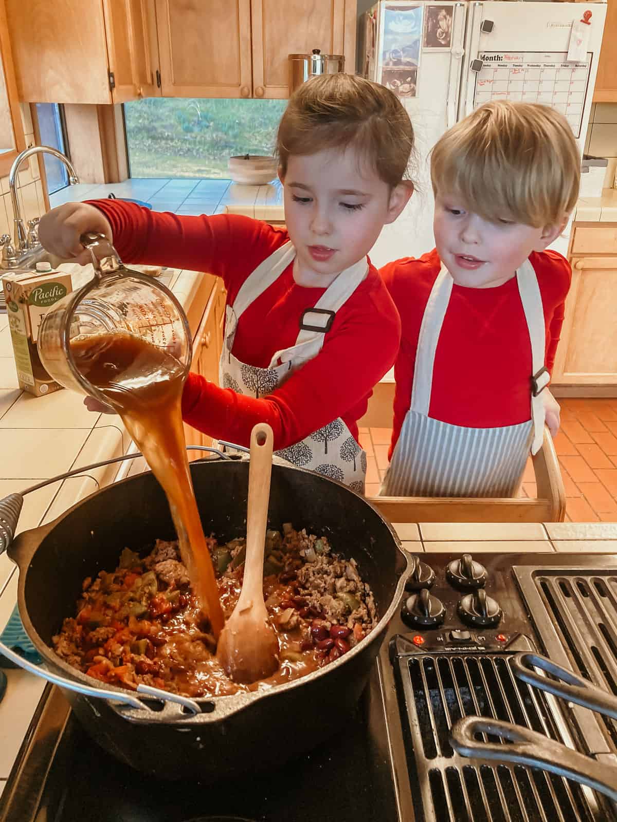 kids in a learning tower. One child is pouring beef stock into a dutch oven and another child is watching