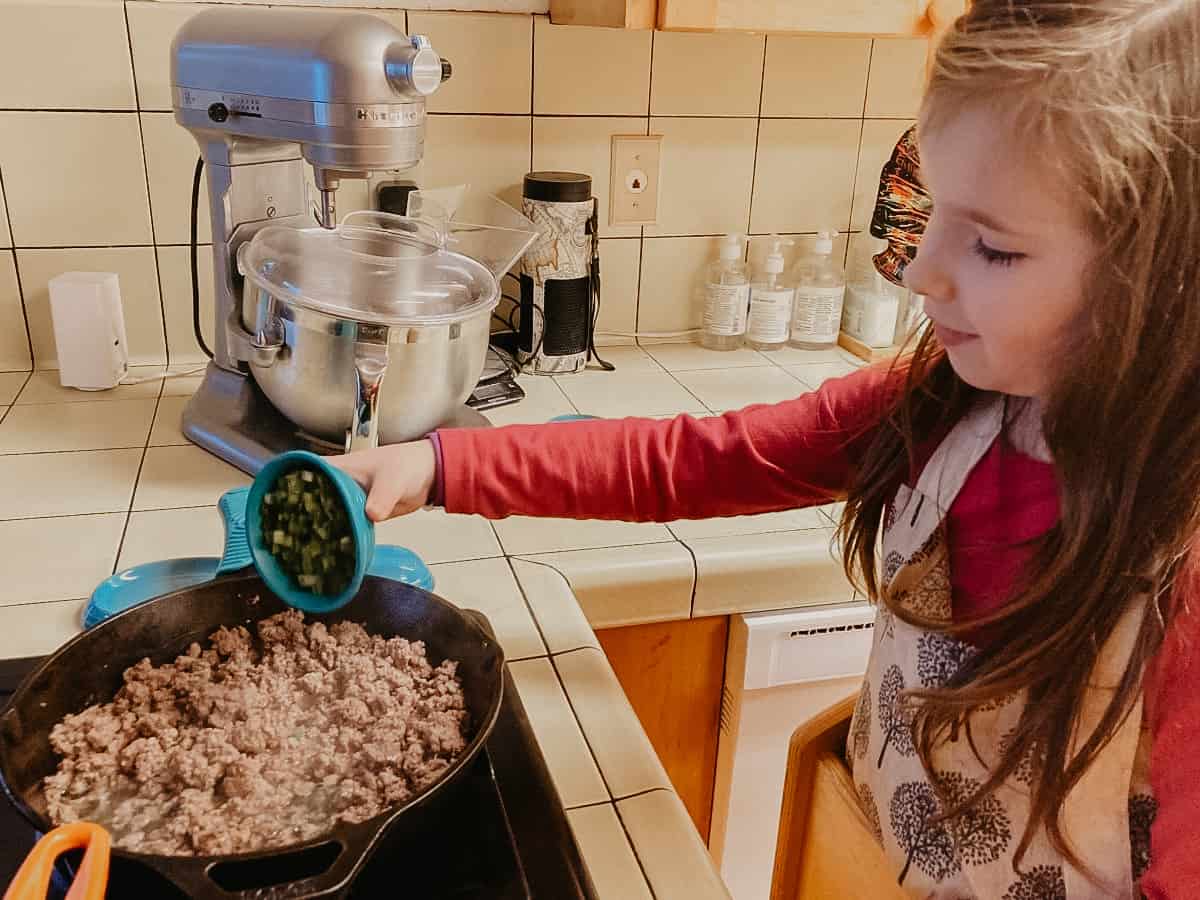 a child adding vegetables to a pan of cooked ground beef