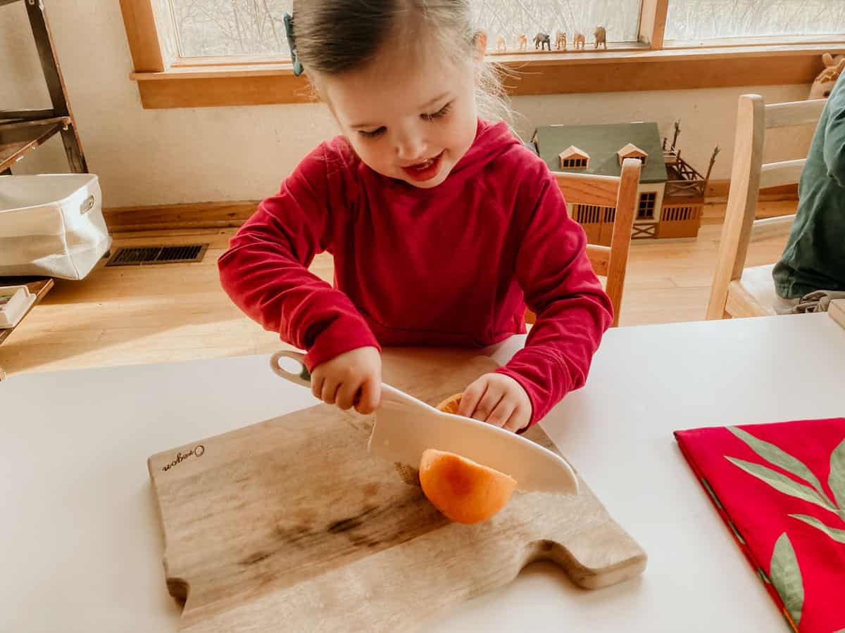 child slicing an orange in half with a child-friendly knife and cutting board