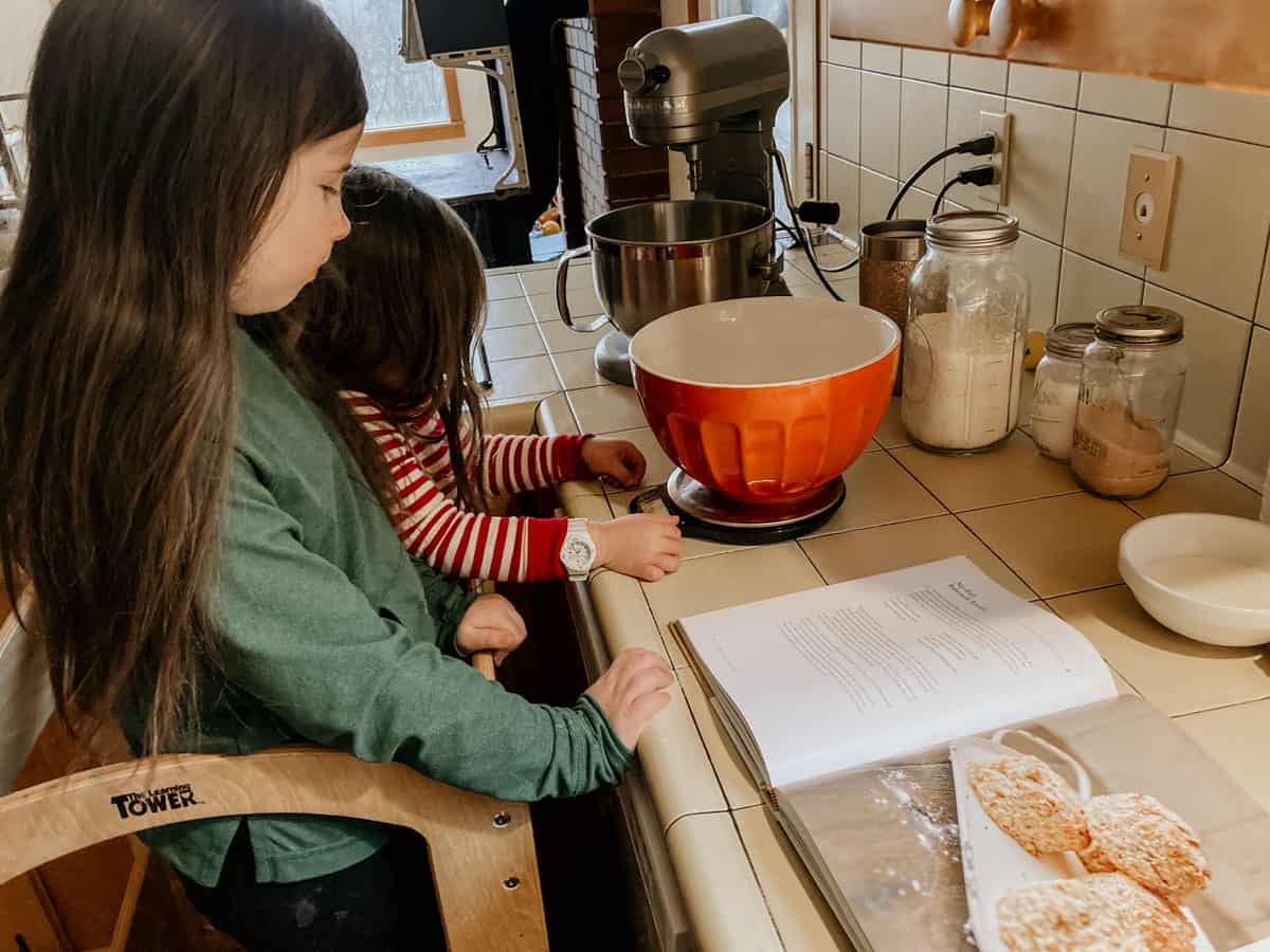 kids in a learning tower at the kitchen counter. One child is reading a digital kitchen scale and another child is watching
