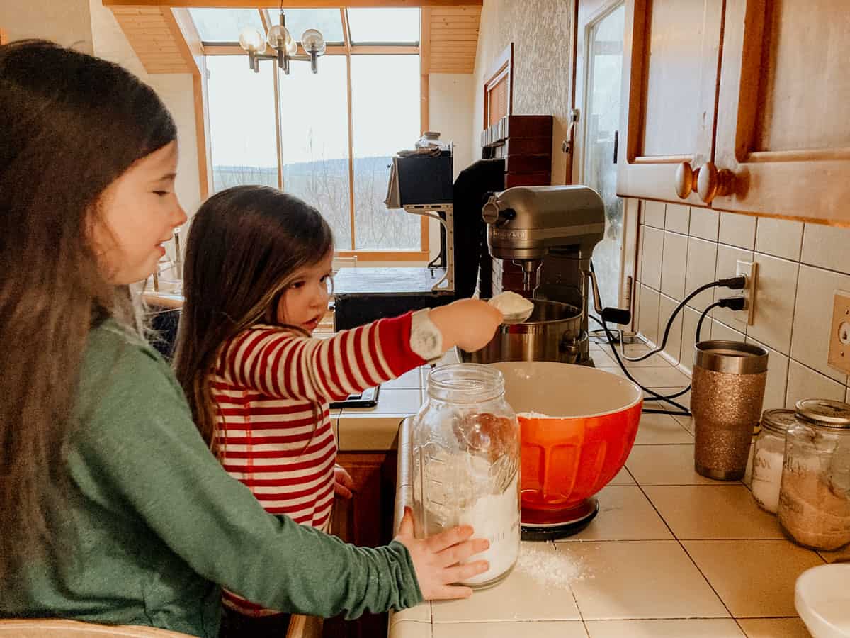 a child is transferring flour to a large mixing bowl and another child is supporting the jar of flour