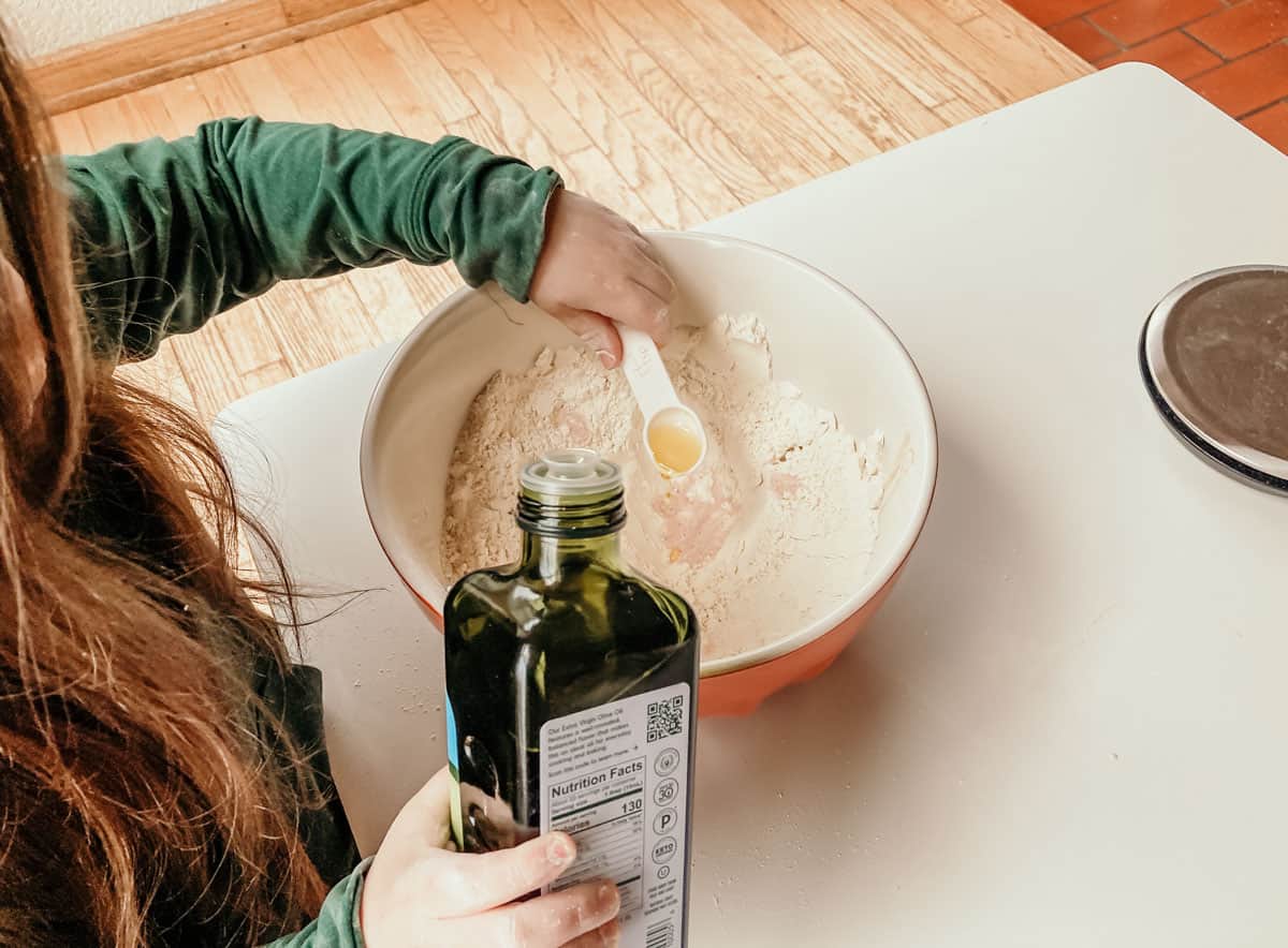 child using a measuring spoon to add olive oil to a bowl of ingredients