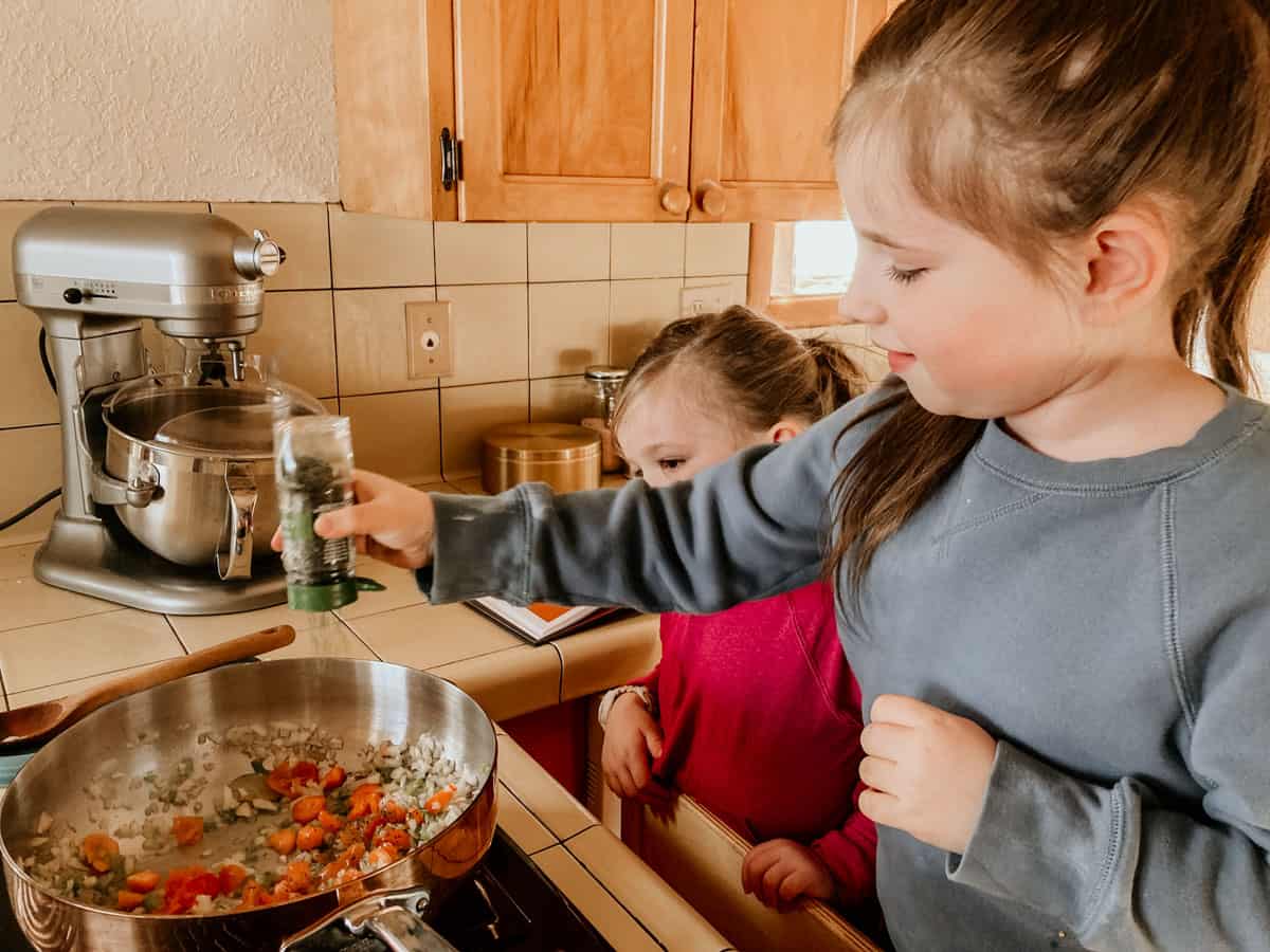 a child at the stove is adding seasoning to a copper saucier of ingredients