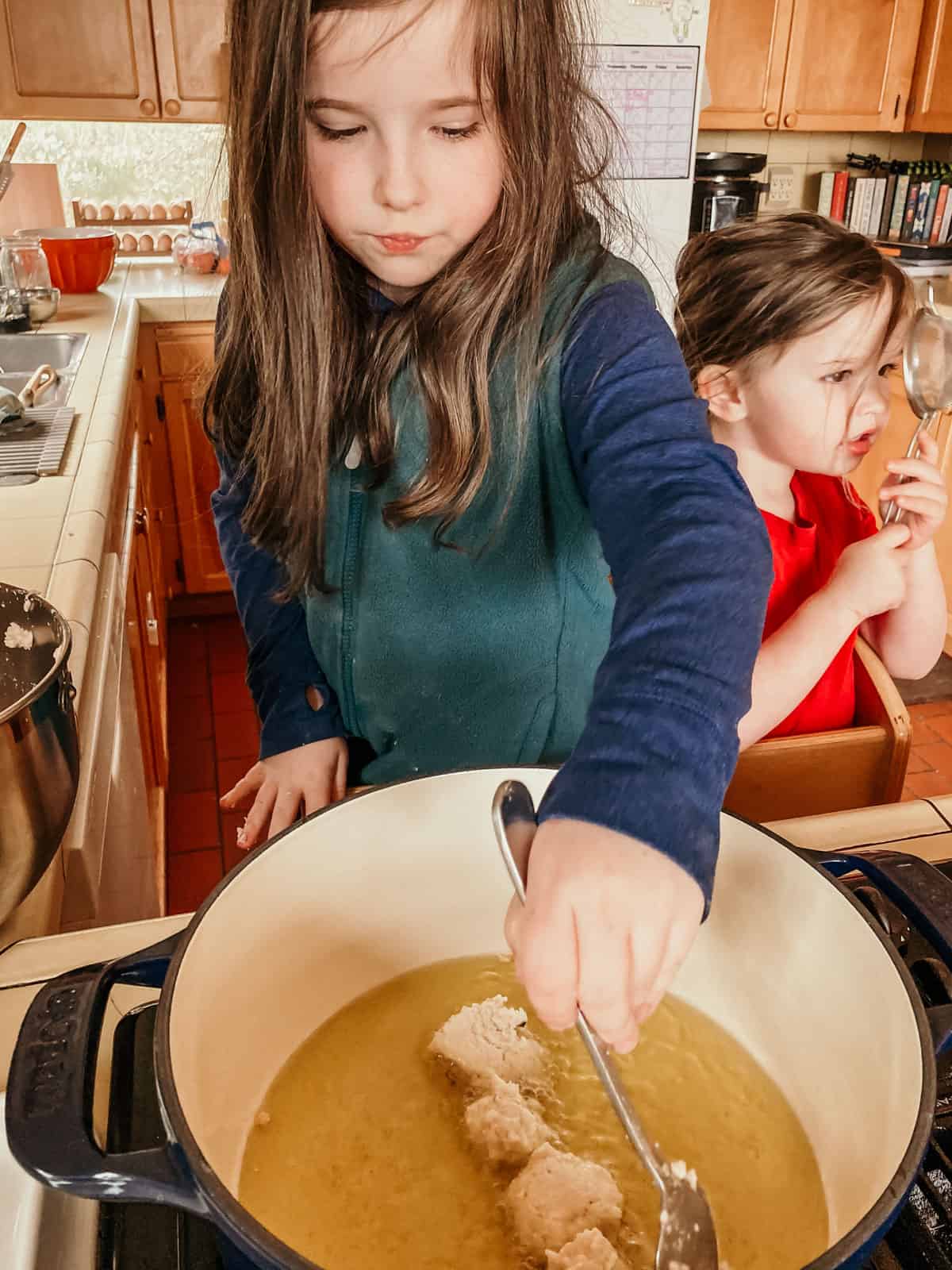 kids in in a learning tower at the stove. One child is placing black-eyed pea puffs in a dutch-oven and the other child is looking through a fine mesh strainer
