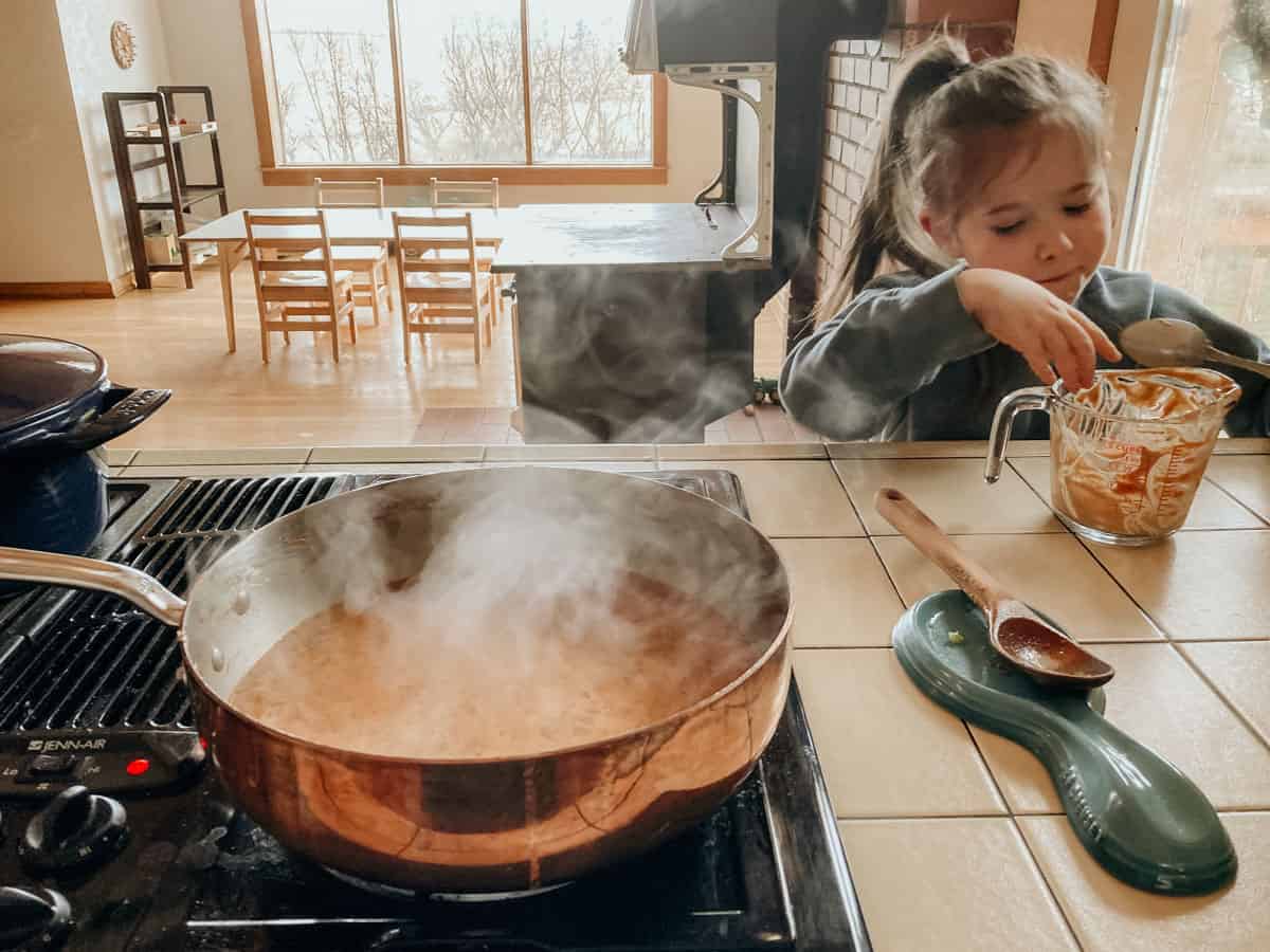 A Made In Copper Saucier on a kitchen stove. The Mother Africa Sauce inside is simmering. A child is next to the stove eating peanut butter out of a measuring cup.