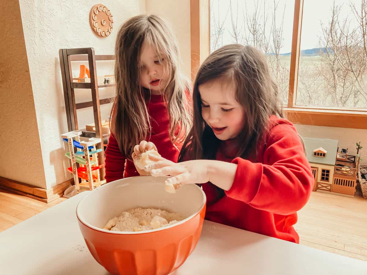 one child is combining flour and butter in a large bowl and another child is watching