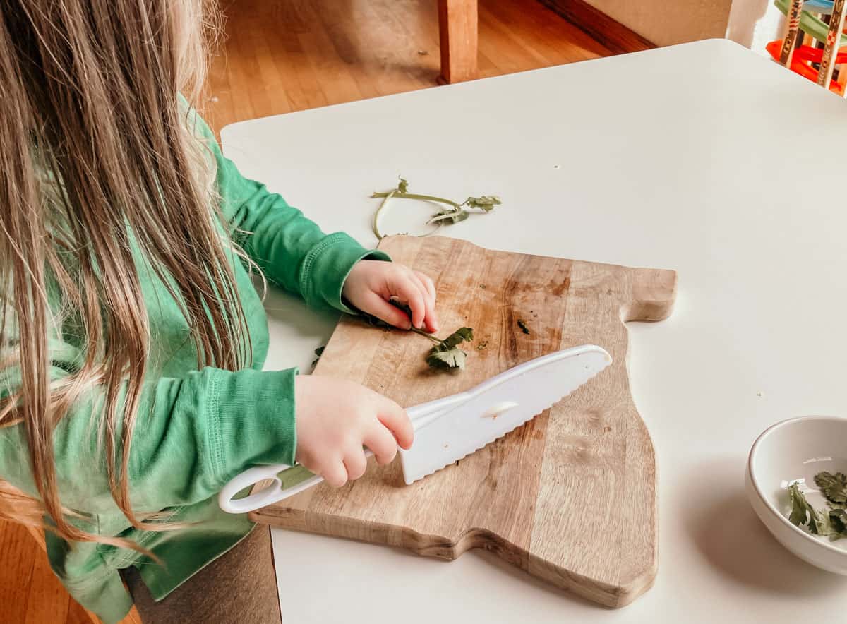 child holding cilantro and a knife over a cutting board