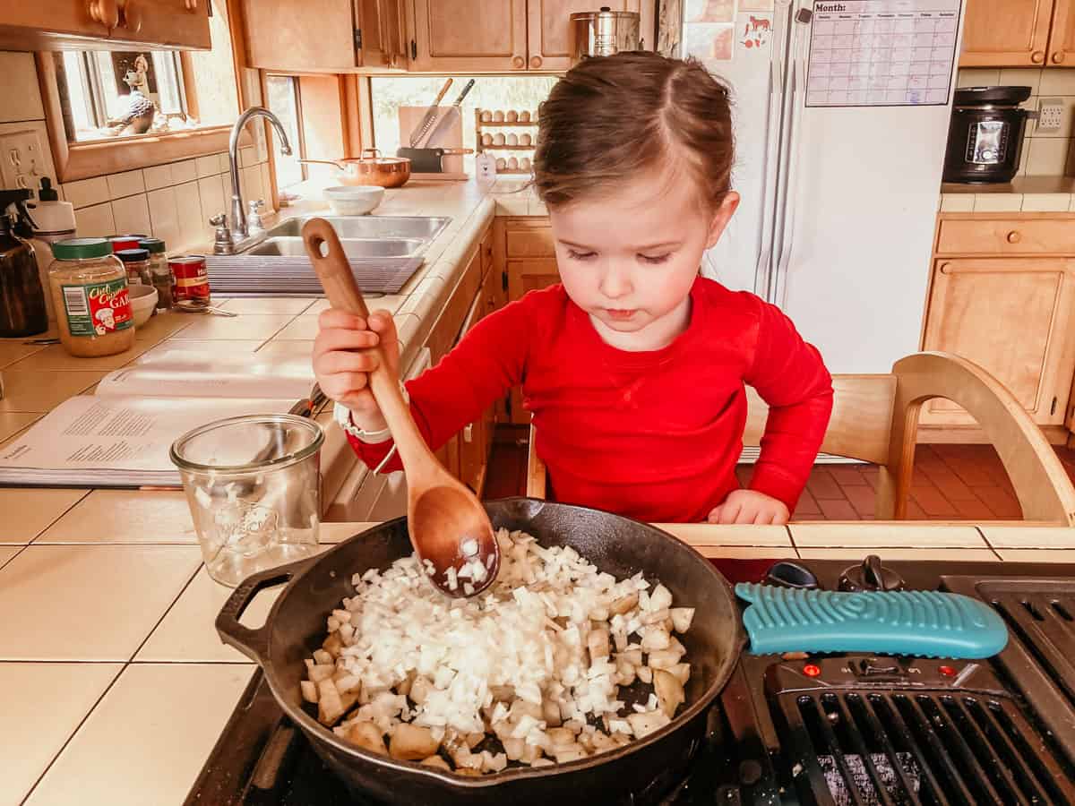 child standing in a learning tower at the stove and combining ingredients in a pan