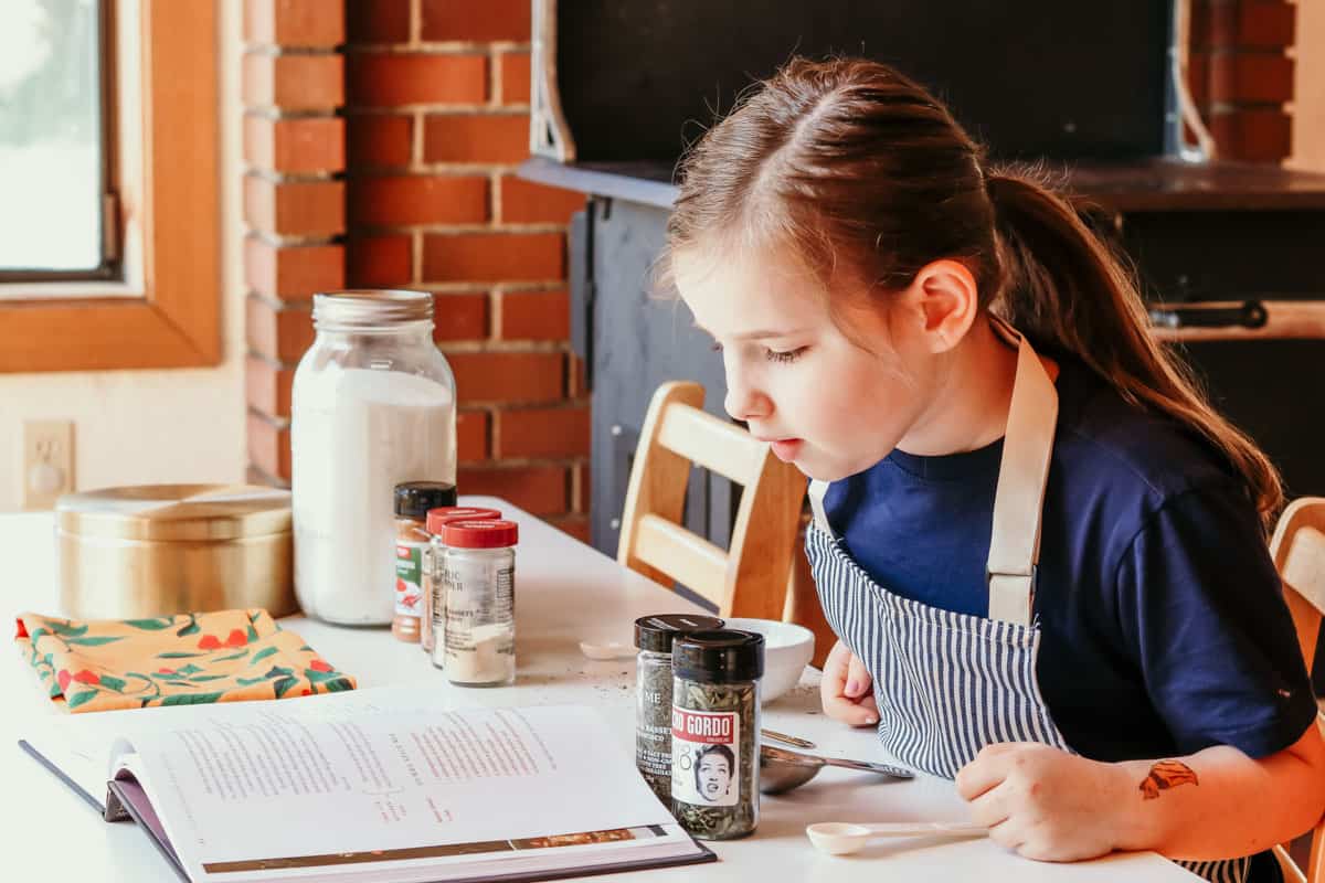 a child sitting at a table with spices and reading a recipe from Between Harlem and Heaven cookbook for making a gumbo spice mix