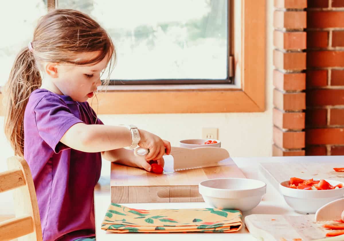 a child sitting at a table slicing grape tomatoes with a child-friendly knife and butcher block