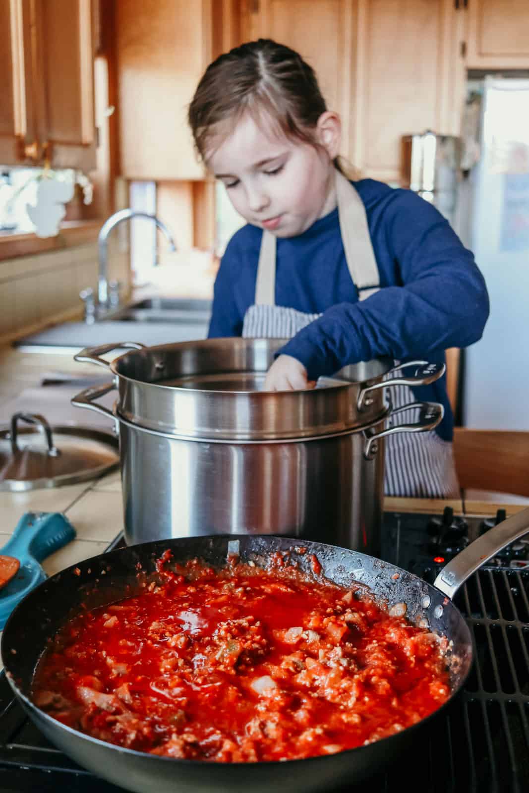 pasta sauce cooking in a carbon steel pan in front of a stock pot with pasta insert. a child is standing over the stock pot and reaching in