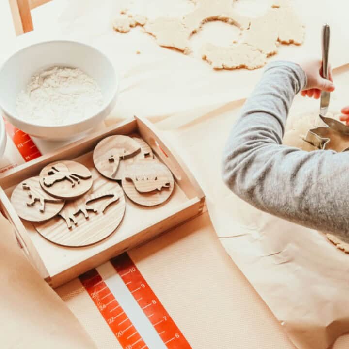 Kaia is using a knife to cut the dough away from the dinosaur cookie cutter