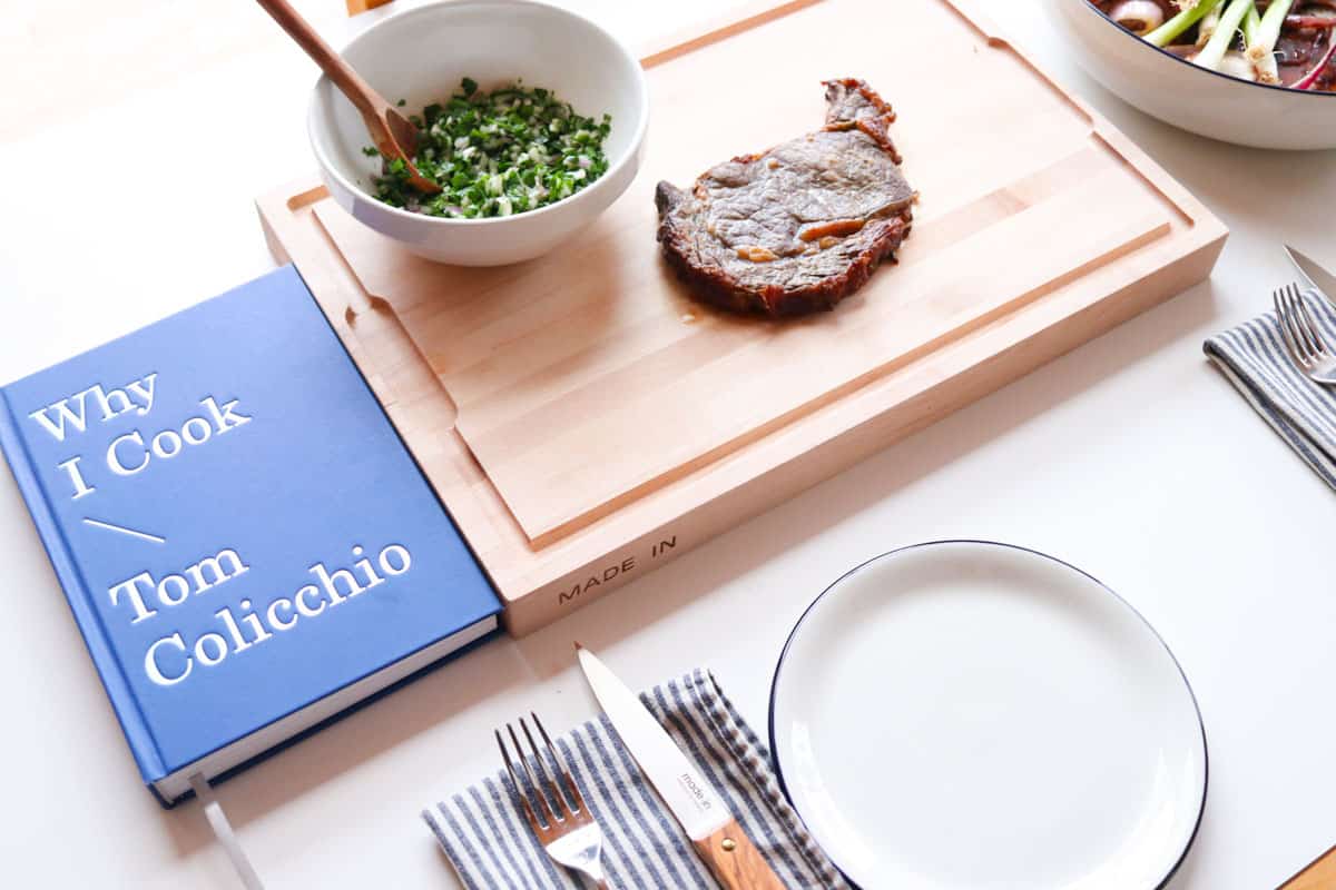 Why I Cook, salsa verde, and rib eye on a butcher block beside place settings