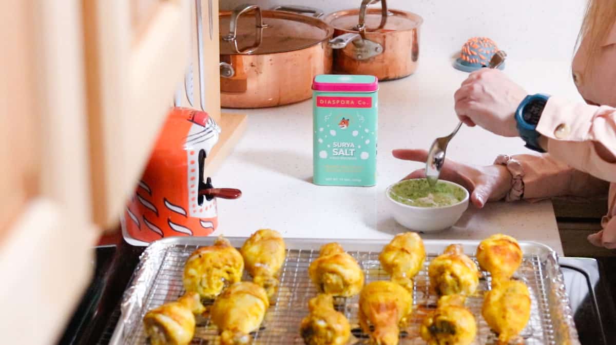 Kristin is mixing cilantro chutney with Greek yogurt beside a tray of tandoori chicken drumsticks