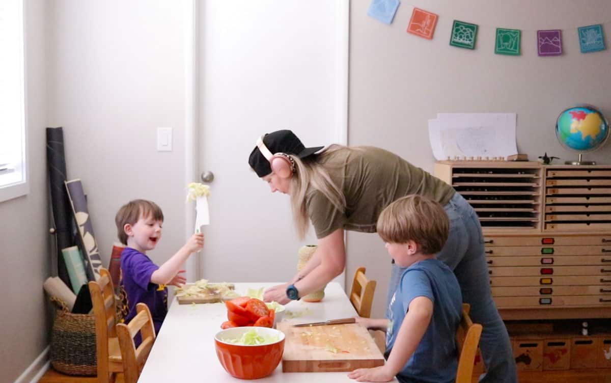 John is holding up cabbage on his knife and Steven is helping Kristin clean up