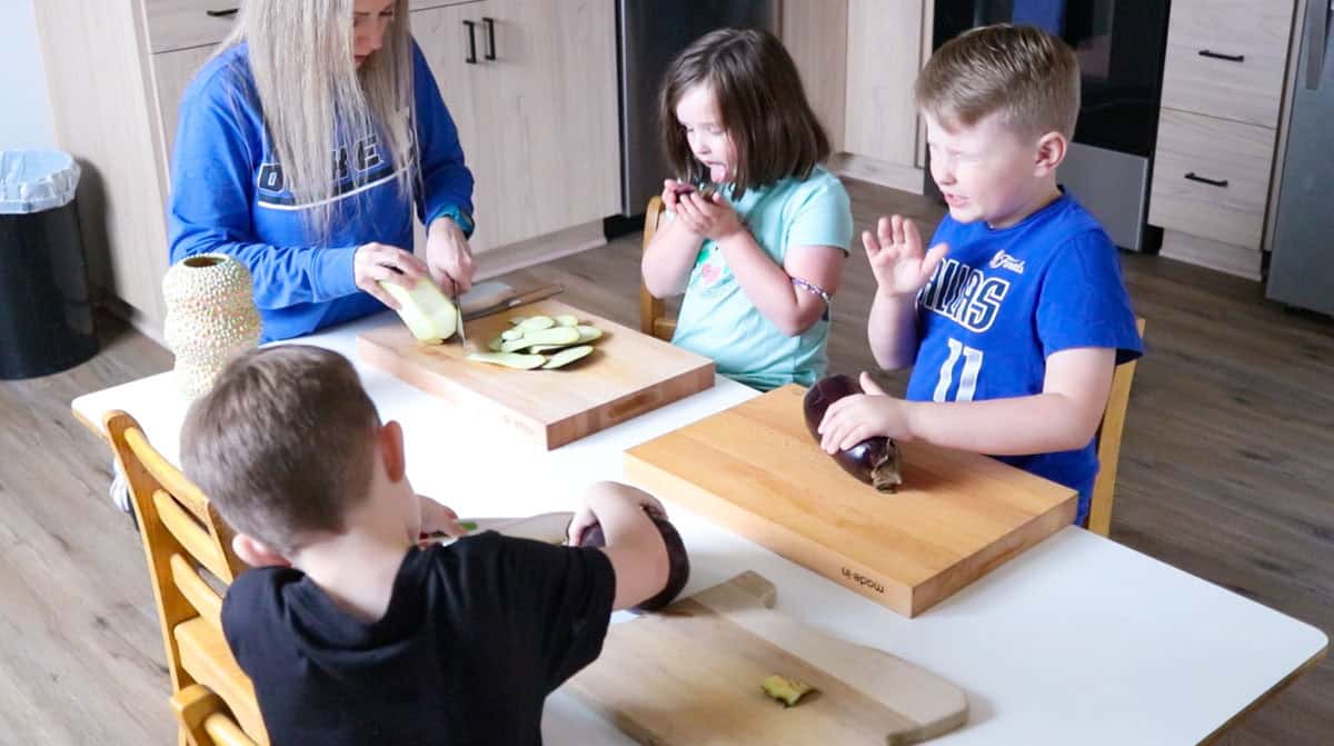 Kristin is showing the kids how to cut the skin off the eggplant. Katalina is licking the eggplant and Noah is grossed out. John is working on his eggplant.