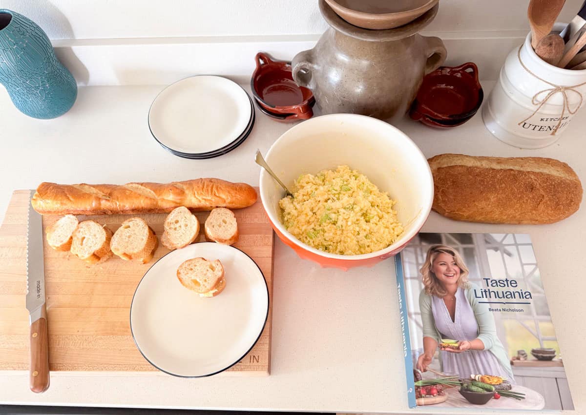 egg salad in a bowl beside a cutting board with bread, a bread knife, and Taste Lithuania cookbook