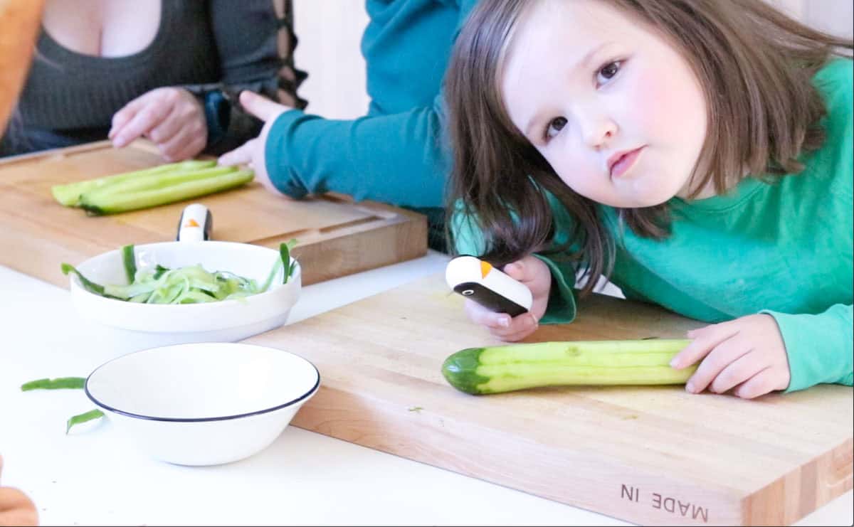 Katalina is looking at the camera while peeling a cucumber and Kristin is showing Kaia how to remove the seeds