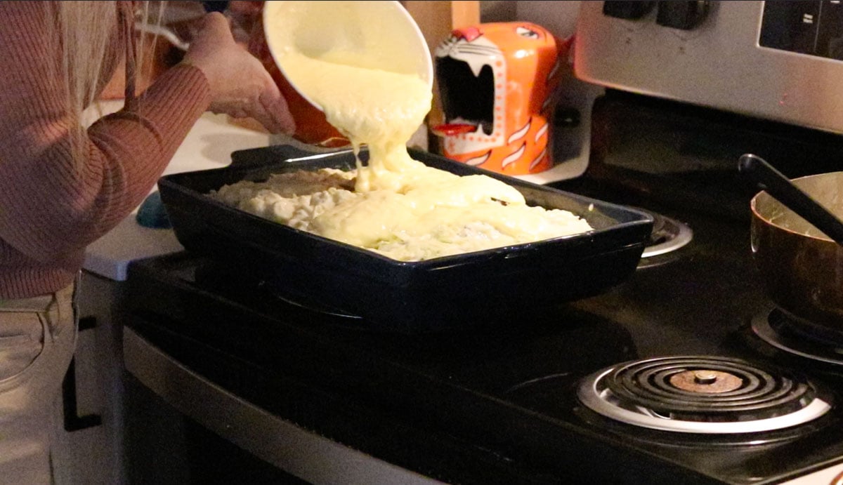 egg and yogurt mixture being poured over musaka ingredients in a baking dish