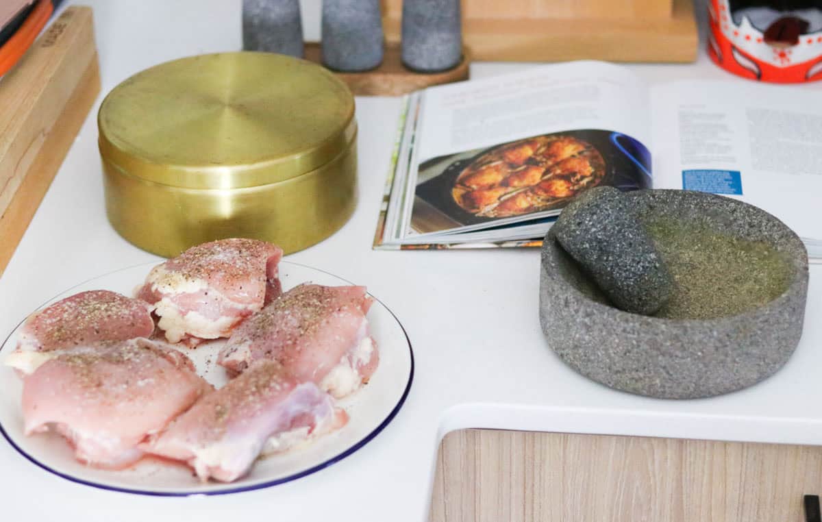 raw seasoned chicken thighs on a plate beside Does This Taste Funny? cookbook and a masala dabba