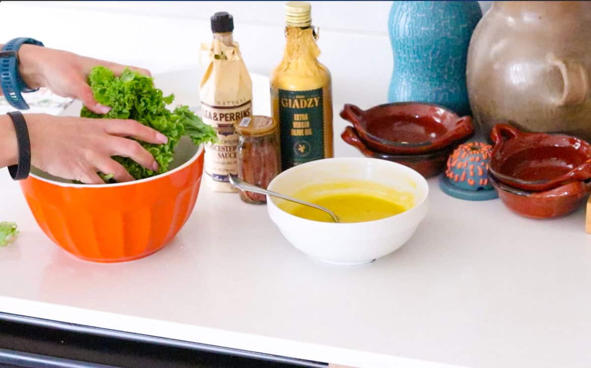 Caesar dressing in a bowl beside giadzy olive oil, worcestershire sauce, anchovies, and Kristin massaging some very lucky kale