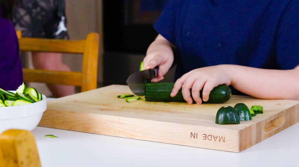 Noah is slicing a zucchini into thin slices using a chef knife and butcher knife