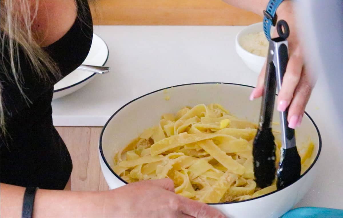 Kristin is using tongs to coat the pappardelle pasta in butter and parmesan