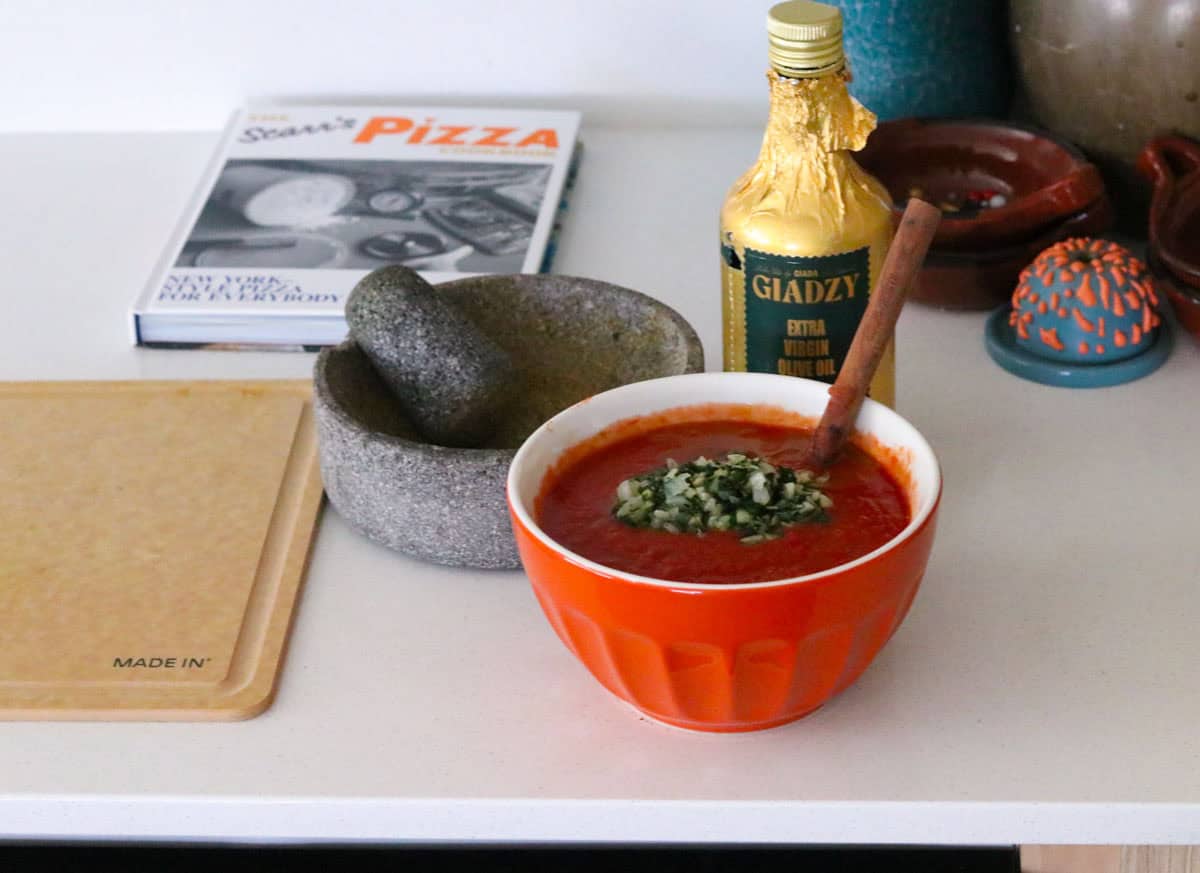 cooked onion, garlic, oregano, and basil in a tomato mixture beside a molcajete with fresh black pepper, a bottle of Giadzy olive oil, a cutting board, and The Scarr's Pizza cookbook