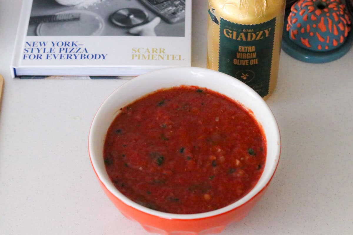 a bowl of ny pizza sauce beside The Scarr's Pizza Cookbook and a bottle of Giadzy extra virgin olive oil
