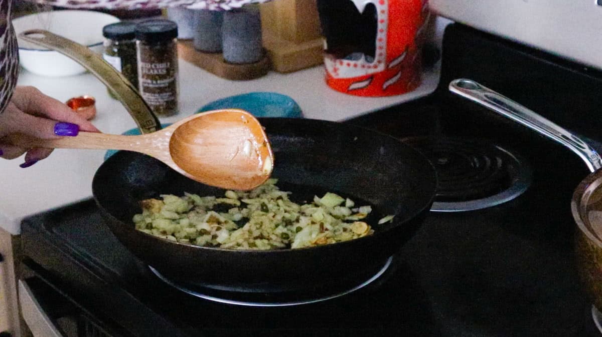 Kristin is stirring garlic, onion, and capers in a pan